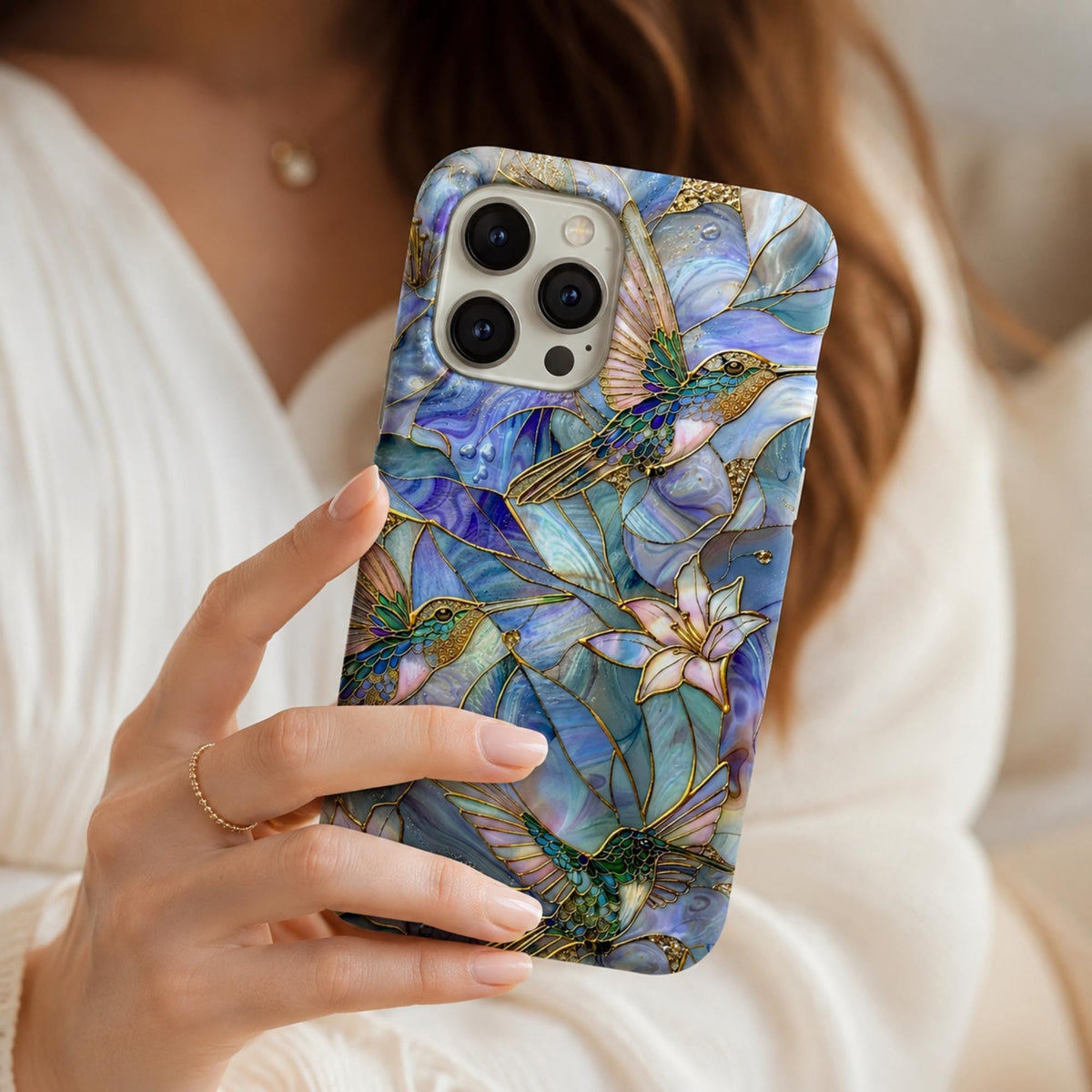 Woman holding a stained glass effect iPhone case featuring vibrant hummingbirds, white lilies, and botanical artwork in shades of blue, teal, ivory, and gold. The glossy protective case showcases a smooth printed stained glass-inspired design with elegant gold accents and edge-to-edge artwork. Captured in soft natural light with a clean neutral background to highlight the premium artistic design. Available from TrioAuraDesign.