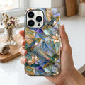 Hand holding a stained glass effect iPhone case featuring colorful hummingbirds, white lilies, and botanical artwork in shades of blue, teal, ivory, and gold. The smooth glossy case showcases a printed stained glass-inspired design with vibrant details and premium edge-to-edge printing. Displayed in soft natural daylight inside a bright home setting, highlighting the elegant artwork and protective design. Available from TrioAuraDesign.