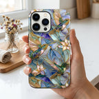 Hand holding a stained glass effect iPhone case featuring colorful hummingbirds, white lilies, and botanical artwork in shades of blue, teal, ivory, and gold. The smooth glossy case showcases a printed stained glass-inspired design with vibrant details and premium edge-to-edge printing. Displayed in soft natural daylight inside a bright home setting, highlighting the elegant artwork and protective design. Available from TrioAuraDesign.