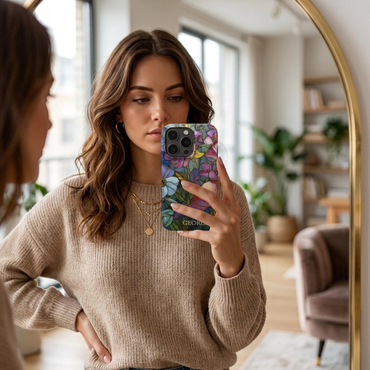 Woman taking a mirror selfie with a personalized iPhone case featuring the custom name Georgia and a colorful stained glass effect in pink, purple, blue, green and golden tones. The lifestyle view shows the botanical artwork and personalization during everyday use, with layered flowers and intricate printed outlines wrapping across the case. The stained glass appearance is a printed visual effect on a smooth glossy Tough or MagSafe Tough case from TrioAuraDesign.