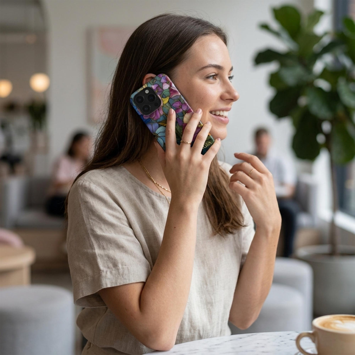 Woman using an iPhone case with a colorful stained glass effect featuring layered pink, purple and blue flowers, green leaves and intricate golden-looking printed outlines. The lifestyle café scene shows how the botanical case looks during everyday use while highlighting the full-wrap artwork and protective camera area. The stained glass appearance is a printed visual effect on a smooth glossy Tough or MagSafe Tough case from TrioAuraDesign.