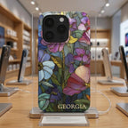 Personalized iPhone case with the custom name Georgia displayed upright in a modern phone store, featuring a colorful stained glass effect with layered pink, purple, blue, green and yellow botanical artwork and intricate golden-looking printed outlines. The product-focused view clearly shows the full-wrap design, personalization and protective camera area. The stained glass appearance is a printed visual effect on a smooth glossy Tough or MagSafe Tough case from TrioAuraDesign.