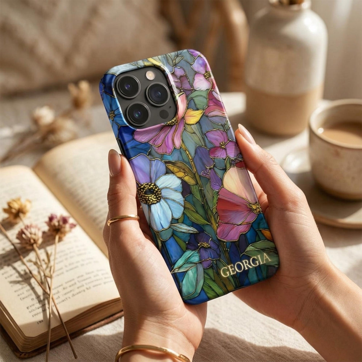 Personalized iPhone case with the custom name Georgia, featuring a colorful stained glass effect with layered pink, purple, blue and coral flowers, green leaves and intricate golden-looking printed details. Held in both hands in a warm neutral setting, the close-up view highlights the vibrant botanical artwork, personalization and protective camera area. The stained glass appearance is a printed visual effect on a smooth glossy Tough or MagSafe Tough case from TrioAuraDesign.