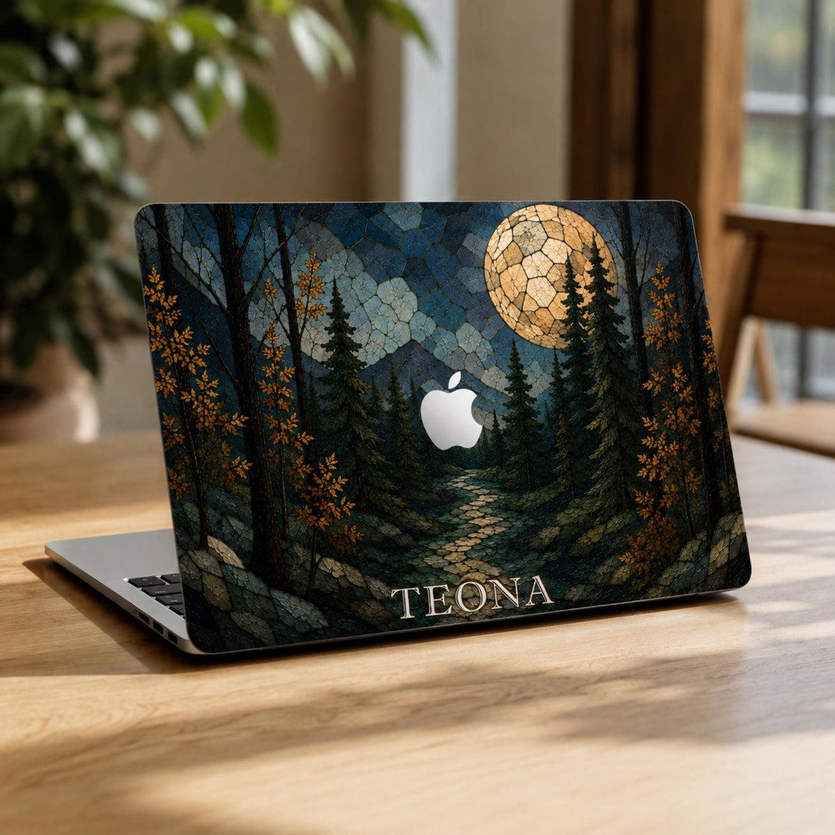 Open MacBook on a wooden table with a personalized stained glass effect hard shell case featuring a dark evergreen forest, golden autumn leaves, blue mountains, winding path and large glowing full moon. The custom name “TEONA” is printed in light serif lettering along the bottom, with the Apple logo visible in the center. Personalized fall laptop cover for MacBook Air 13 15, MacBook Pro 13 14 16 and MacBook Neo by TrioAuraDesign.