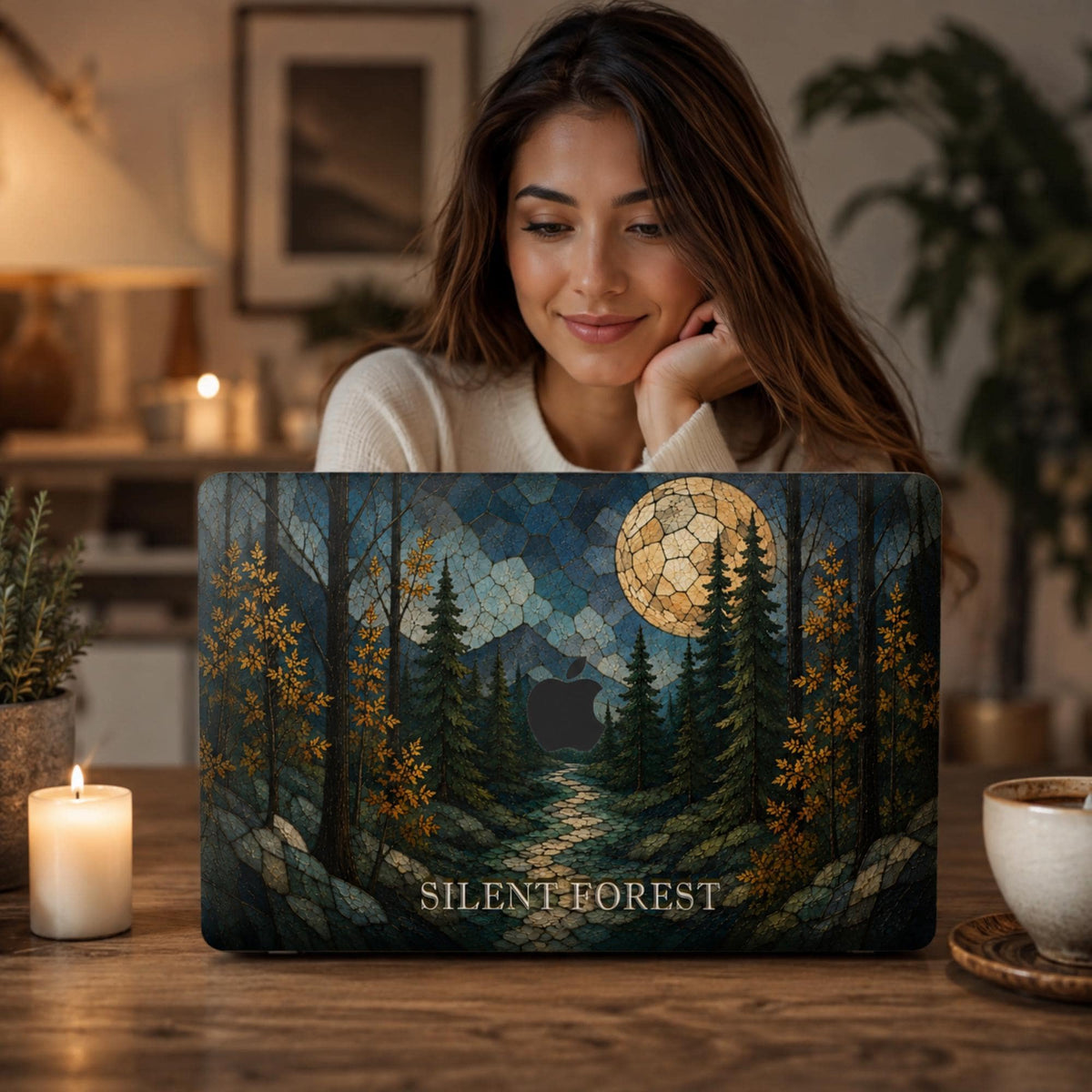 Woman sitting behind a personalized stained glass effect MacBook hard shell case featuring a dark evergreen forest, golden autumn leaves, blue mountains, winding path and glowing full moon. “SILENT FOREST” is printed in light serif lettering along the bottom, with the Apple logo area visible in the center. Custom fall laptop cover for MacBook Air 13 15, MacBook Pro 13 14 16 and MacBook Neo. Cozy nature-inspired case by TrioAuraDesign.