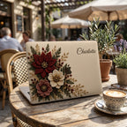 Personalized ivory Crochet effect floral MacBook case from TrioAuraDesign, displayed on a rustic outdoor café table with a decorative coffee cup and potted plants. The smooth hard shell features deep burgundy, ivory and dusty rose flowers with botanical greenery, berries and gold-toned details, custom name “Charlotte” in elegant script, and the Apple logo visible at center. Floral laptop cover for MacBook Air, Pro and Neo models.