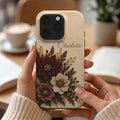 Personalized ivory embroidery effect floral iPhone case from TrioAuraDesign, shown held in both hands in a warm neutral setting. The smooth printed case features deep burgundy, ivory and dusty rose flowers with botanical greenery, berries and gold-toned details. Custom name “Charlotte” appears near the upper right below the camera area. Elegant personalized floral phone case with a realistic embroidered look and smooth finish.