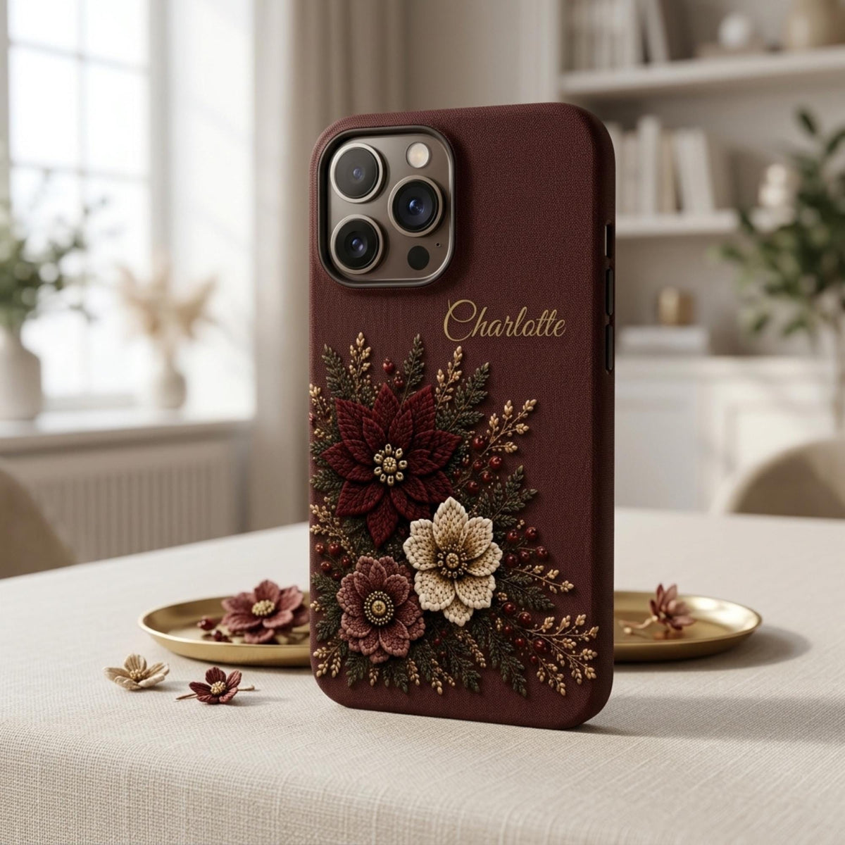 Personalized deep wine embroidery effect floral iPhone case from TrioAuraDesign, displayed upright in a warm neutral interior. The smooth printed phone case features burgundy, ivory and dusty rose flowers with botanical greenery, berries and gold-toned details, plus the custom name “Charlotte” near the upper right. Elegant personalized floral iPhone case with a realistic embroidered look and smooth finish, available in my shop.