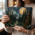 Woman holding a personalized MacBook hard shell case featuring a stained glass effect night forest scene with dark evergreen trees, golden autumn leaves, blue mountains, a winding path and a large glowing full moon. The words “SILENT FOREST” are printed in large light-colored serif letters along the bottom of the case, with the Apple logo visible near the center. Cozy fall lifestyle mockup by TrioAuraDesign.