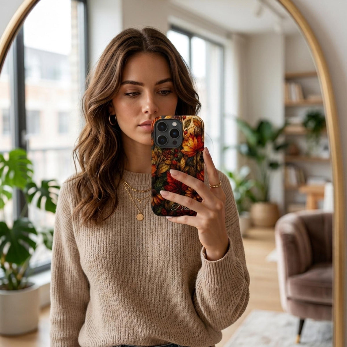 Personalized autumn stained glass effect iPhone case from TrioAuraDesign, shown in a mirror selfie in a bright modern home. The smooth printed case features rich maple leaves, berries and florals in burnt orange, deep red, golden yellow and olive tones, creating a stained glass mosaic look. Custom name personalization appears near the bottom, adding an artistic fall finish available in my shop.