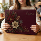 Personalized deep wine Crochet effect floral MacBook case from TrioAuraDesign, shown being held in a cozy café setting. The smooth hard shell features rich burgundy, ivory and dusty rose flowers with botanical greenery, berries and gold-toned details. Custom name “Charlotte” appears in elegant script at the upper right, with the Apple logo visible at center. Floral laptop cover for MacBook Air, Pro and Neo models, available in my shop.