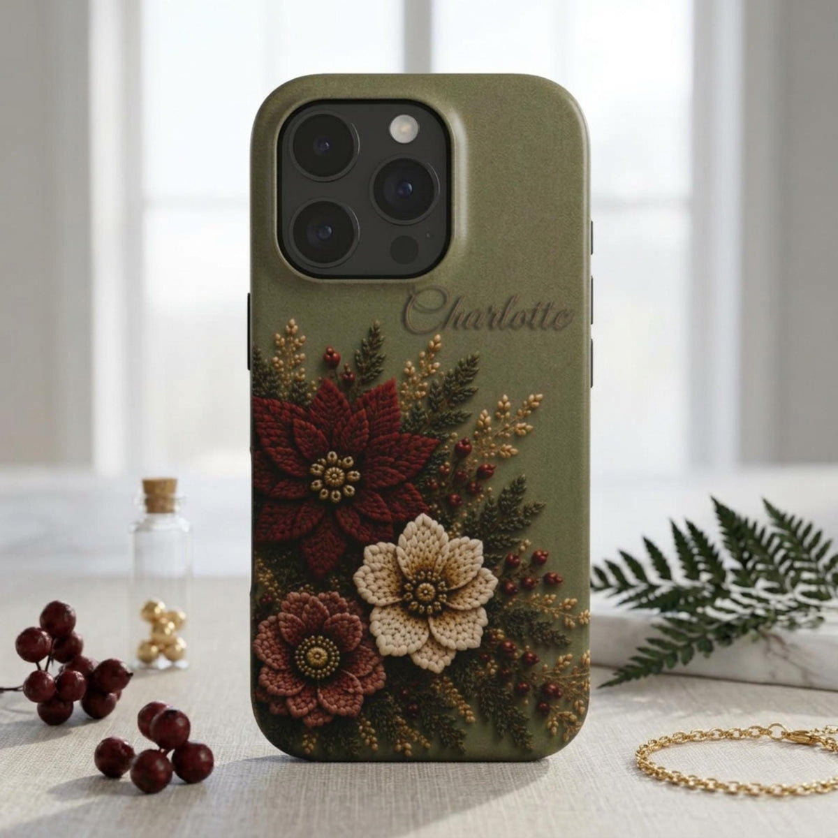 Personalized sage green embroidery effect floral iPhone case from TrioAuraDesign, displayed upright in a bright neutral setting. The smooth printed case features deep burgundy, ivory and dusty rose flowers with botanical greenery, berries and gold-toned details, plus the custom name “Charlotte” near the upper right. Elegant personalized floral phone case with a realistic embroidered look and smooth finish, available in my shop.
