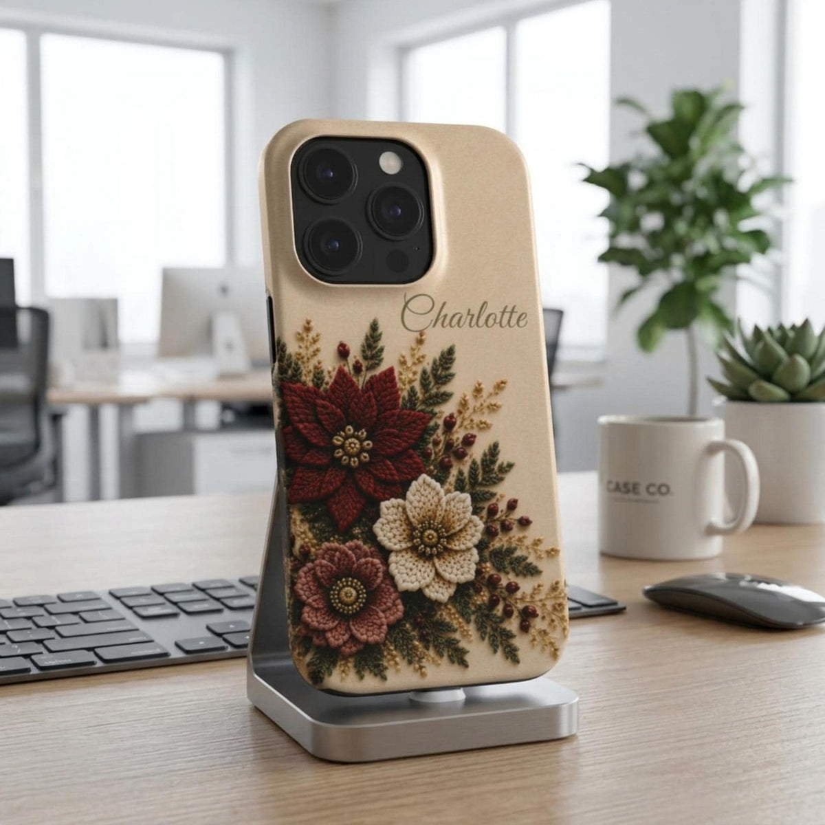 Personalized ivory embroidery effect floral iPhone case from TrioAuraDesign, displayed upright on a silver phone stand in a bright modern workspace. The smooth printed case features deep burgundy, ivory and dusty rose flowers with botanical greenery, berries and gold-toned details, plus the custom name “Charlotte” near the upper right. Elegant personalized floral phone case with a realistic embroidered look and smooth finish, available in my shop.