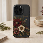 Personalized charcoal embroidery effect floral iPhone case from TrioAuraDesign, displayed upright in a warm neutral setting. The smooth printed case features deep burgundy, ivory and dusty rose flowers with botanical greenery, berries and gold-toned details, plus the custom name “Charlotte” near the upper right. Elegant personalized floral phone case with a realistic embroidered look and smooth finish, available in my shop.
