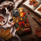 Personalized autumn stained glass effect iPhone case from TrioAuraDesign, styled on a dark wood desk with headphones, an open notebook and scattered fall leaves. The smooth printed case features a rich mosaic of glowing maple leaves, berries and florals in burnt orange, deep red, golden yellow and olive tones, with custom name “Diana” near the bottom. Artistic fall phone case inspired by stained glass and autumn foliage, available in my shop.