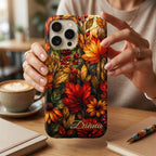 Personalized autumn stained glass effect iPhone case from TrioAuraDesign, shown held upright at a cozy café table beside a latte and notebooks. The smooth printed case features glowing maple leaves, berries and florals in burnt orange, deep red, golden yellow and olive tones, creating a rich stained glass mosaic look. Custom name “Diana” appears near the bottom, adding a personal finishing touch.