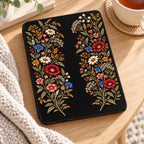 Black Wildflower Kindle Paperwhite case lying on a wooden table beside a cup of tea and a knit blanket, shown without personalization