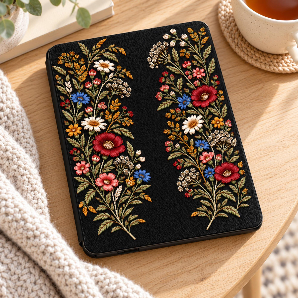 Black Wildflower Kindle Paperwhite case lying on a wooden table beside a cup of tea and a knit blanket, shown without personalization