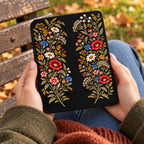 Hands holding a Kindle in a black folio case printed with two garlands of embroidery effect wildflowers, outdoors on an autumn bench