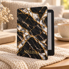 Personalized Black Marble Kindle Paperwhite case in Black Pearl style: black marble effect with gold kintsugi seams and white pearl fragments, with the name ADALINDA printed in gold, standing on a knit blanket