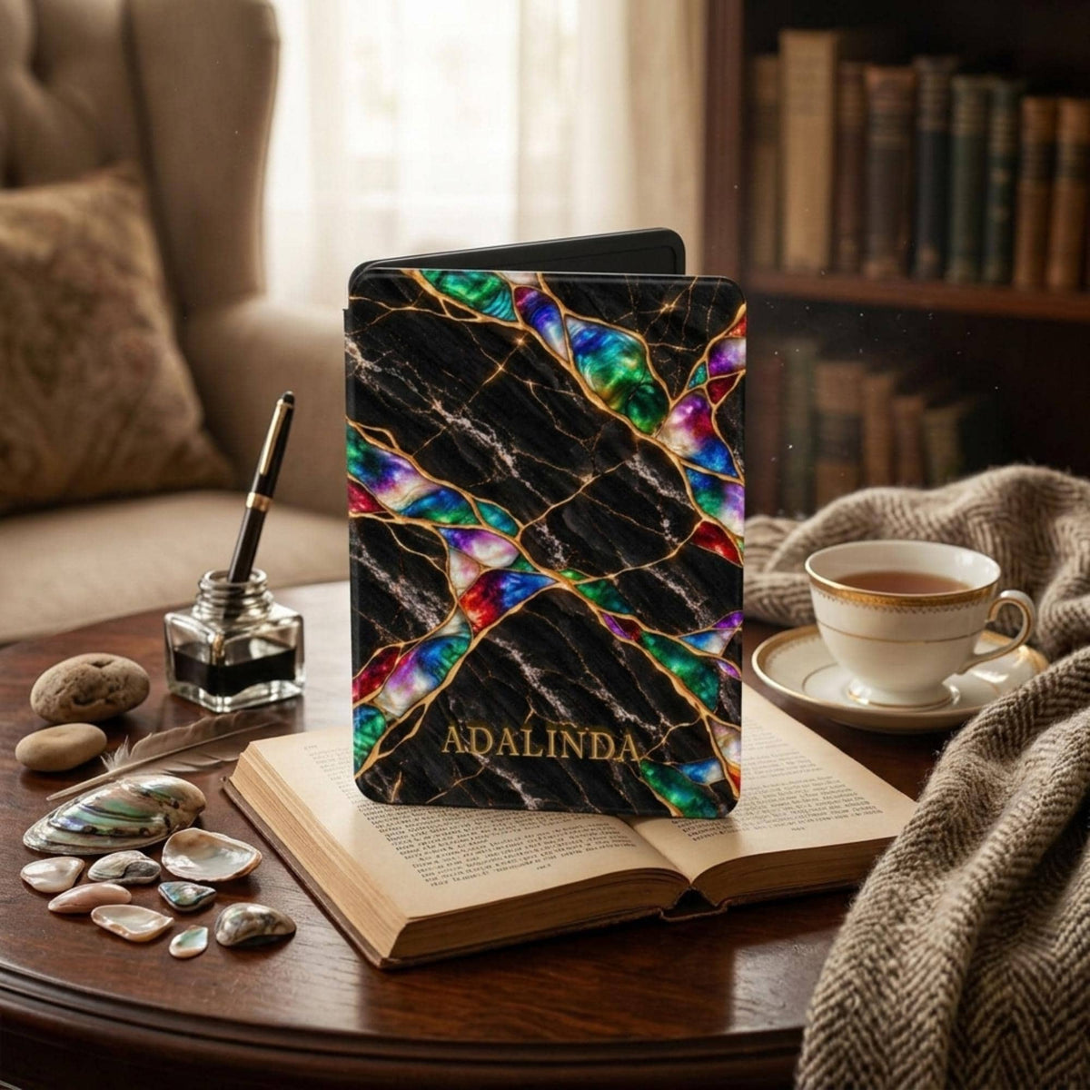 Black Marble Kindle case in Jewel Tone style with gold kintsugi effect seams and emerald, sapphire, violet and ruby opal fragments, personalized with the name ADALINDA in gold, on an open book beside a cup of tea