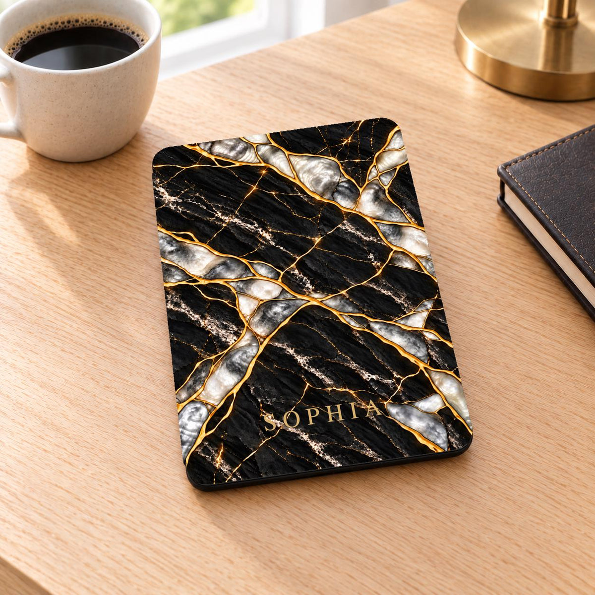 Black Pearl marble Kindle Paperwhite case personalized with the name SOPHIA in gold serif capitals near the bottom of the front cover, lying on a wooden desk next to a coffee cup