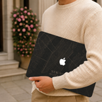 Personalized City Map MacBook case from TrioAuraDesign, shown being carried outdoors with a dark New York street map design. The hard shell features custom city text, coordinates, and the Apple logo visible at center. Close lifestyle view of a meaningful custom map laptop cover for compatible MacBook Air, Pro, and Neo models.