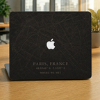 Personalized City Map MacBook case from TrioAuraDesign, shown on a wooden desk with a dark Paris street map design. The smooth hard shell features custom text “PARIS, FRANCE,” coordinates, the phrase “WHERE WE MET,” and the Apple logo visible at center. Custom map laptop cover for compatible MacBook Air, Pro, and Neo models.